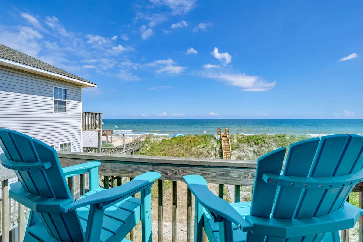 This image captures a tranquil oceanfront deck furnished with turquoise Adirondack chairs, offering stunning panoramic views of the sea and boardwalk. The deck sets the stage for relaxation, al fresco dining, and memorable sunset moments, aligning with the invitation to gather and unwind outdoors.