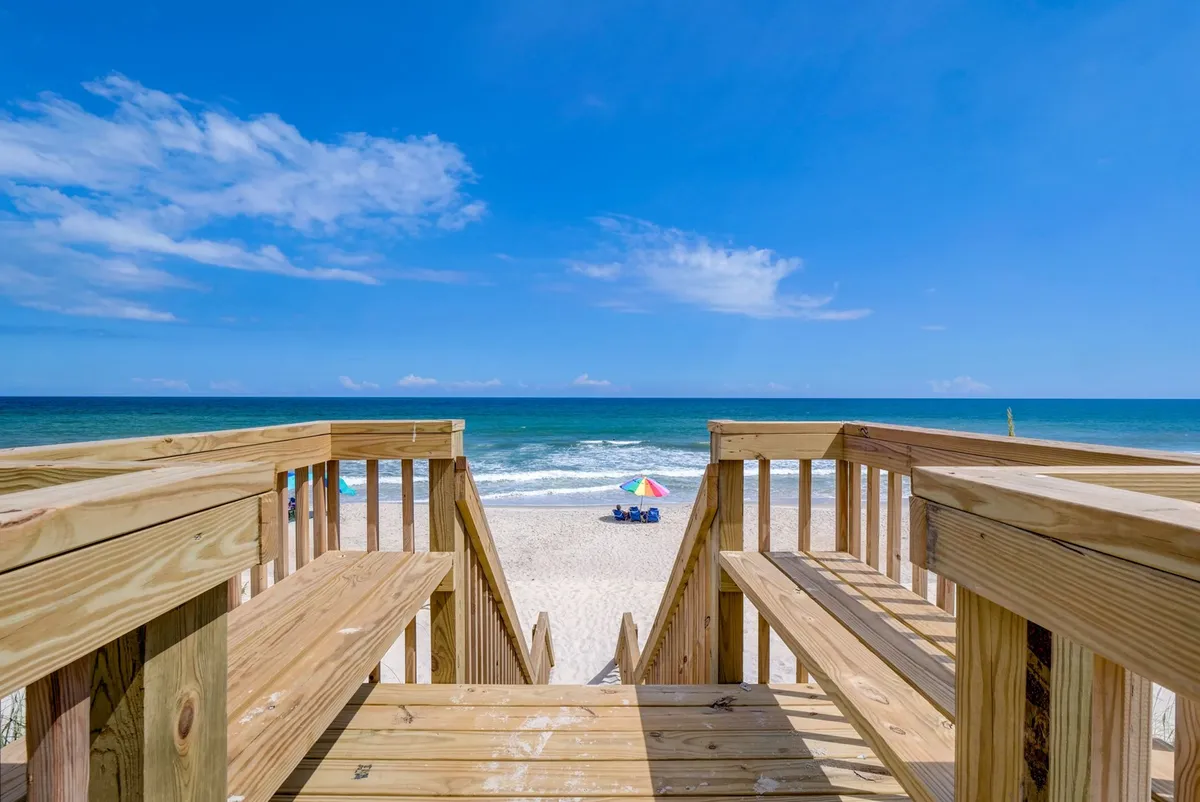 The image captures a scenic exterior view from a wooden staircase leading down to the beach. The ocean stretches out in the background, dotted with colorful beach umbrellas and sunbathers enjoying the sand. The bright sunlight creates a joyful, inviting beach atmosphere that encourages relaxation and play.