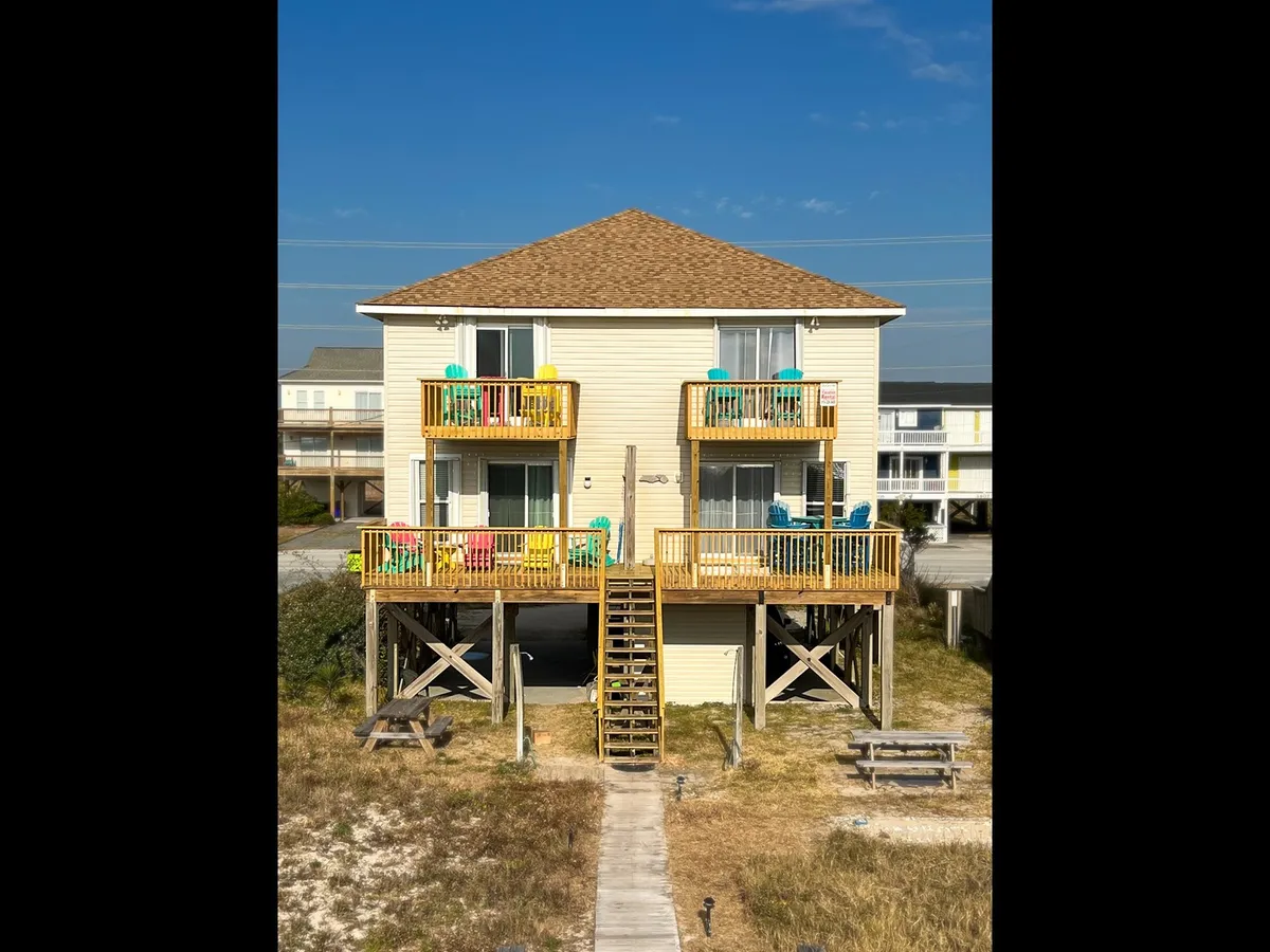 A bright exterior image of a two-story beach house with colorful balconies, a wooden staircase, and an inviting outdoor seating area. The house is clearly designed for groups or couples to relax, entertain, and savor coastal views, embodying both comfort and togetherness.