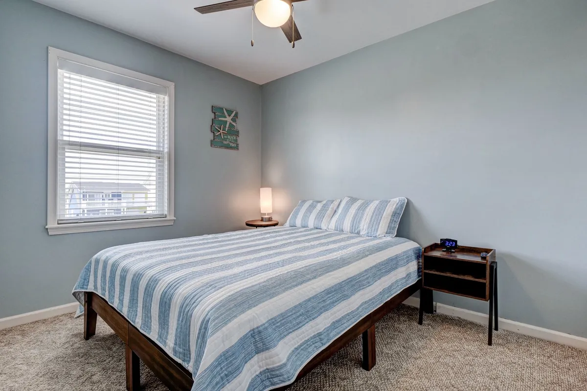 A peaceful queen bedroom featuring a striped bedspread, nightstand with lamp, decorative wall art, and a window letting in gentle sunlight. The setting is serene, designed for restful nights and quiet mornings.