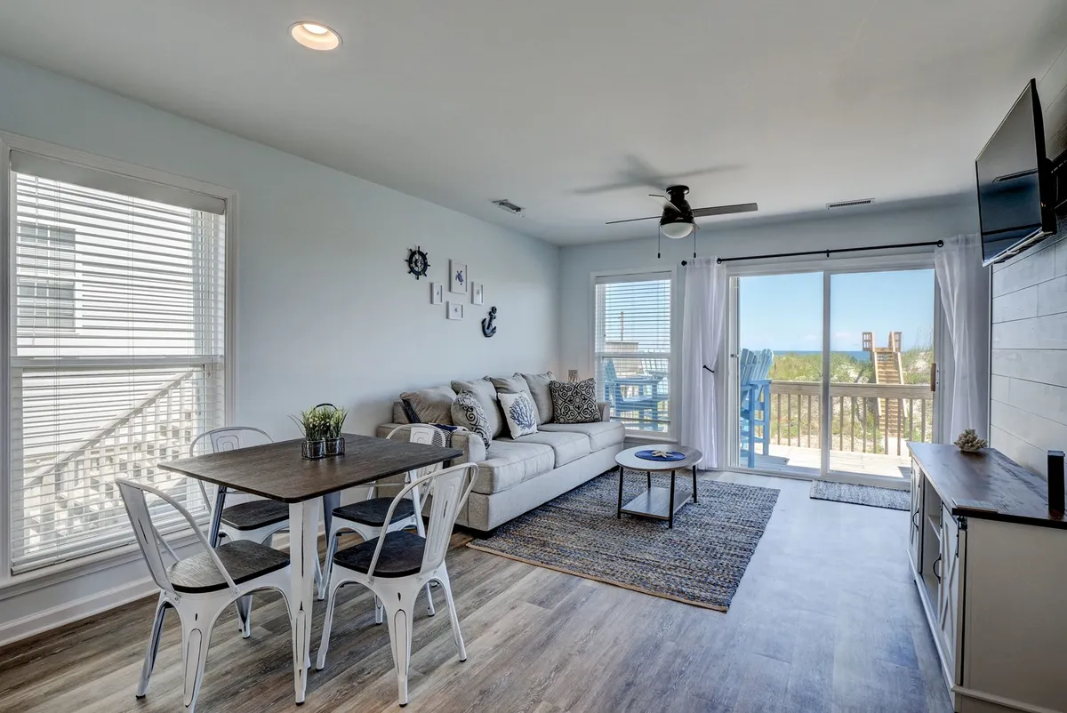 This image presents a cozy living area with a plush sofa, a dining table, and a large sliding glass door leading to a balcony overlooking the ocean. The space is filled with natural light and seamlessly connects the living, dining, and outdoor areas—emphasizing the open-concept layout and the inviting atmosphere ideal for gatherings.