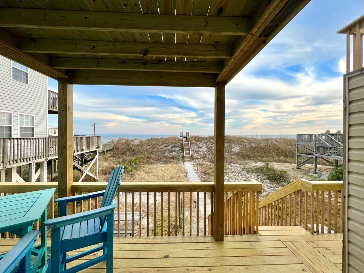 A serene wooden deck with blue adirondack chairs overlooks a path to the beach, bordered by dune grass and open sky. The deck serves as a private, relaxing vantage point from which the beach and ocean can be observed. The space is inviting for parents watching children play or families catching their breath between outings.