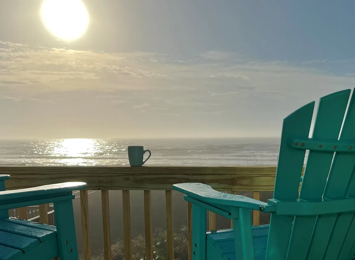 The image features a tranquil exterior view overlooking the ocean at sunrise, with comfortable chairs arranged on a deck and a steaming coffee cup, inviting relaxation. The sun casts a warm glow over the scene, highlighting the proximity to the water and the peaceful ambiance. The gentle breeze and the soothing sound of the waves create a serene and inviting setting for guests.