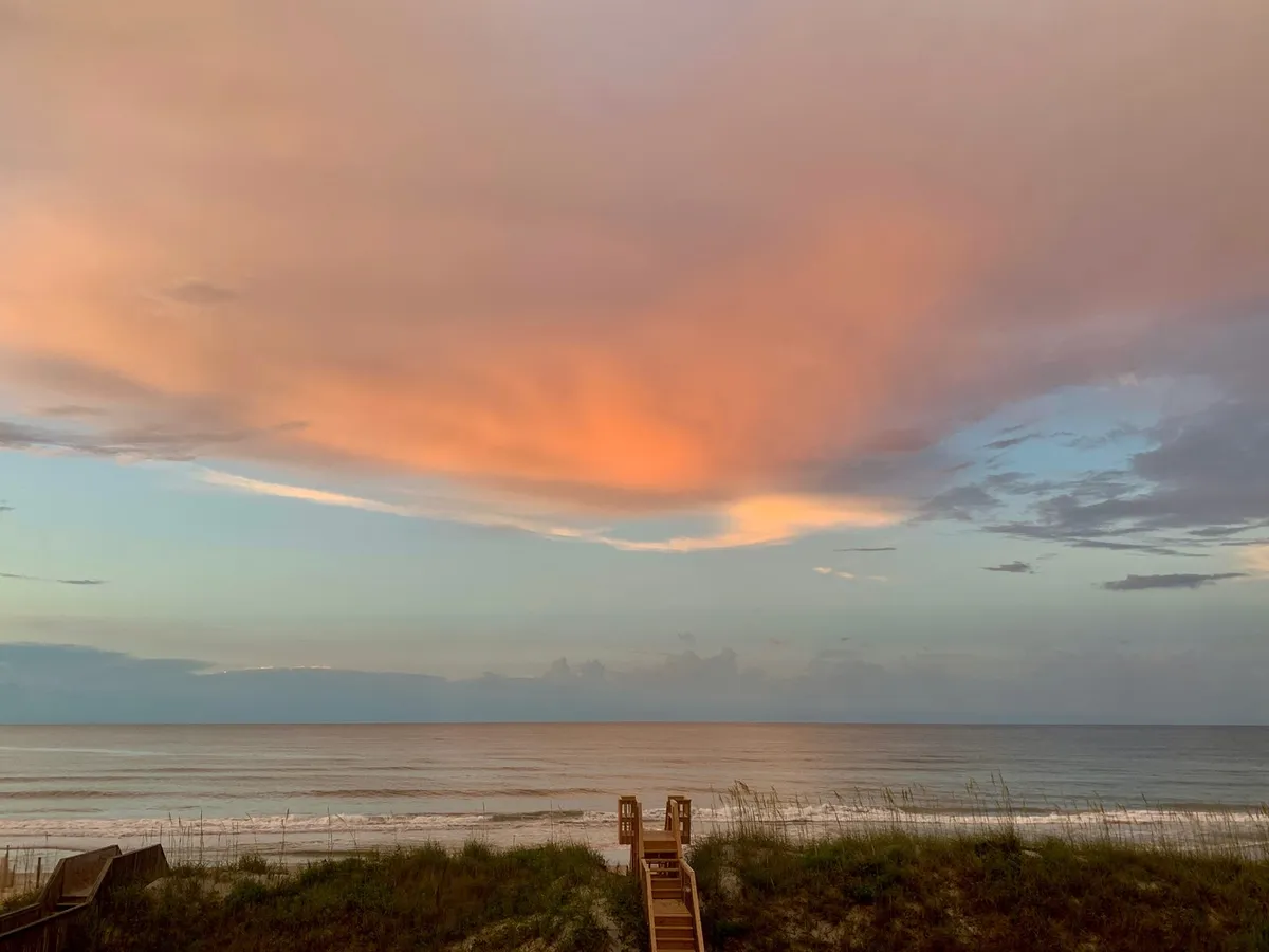 A tranquil exterior shot showing wooden stairs that descend to the sandy shore, with the ocean and colorful sunset sky beyond. The scene feels calm and reflective, perfect for moments of relaxation or inspiration after a day of remote work.