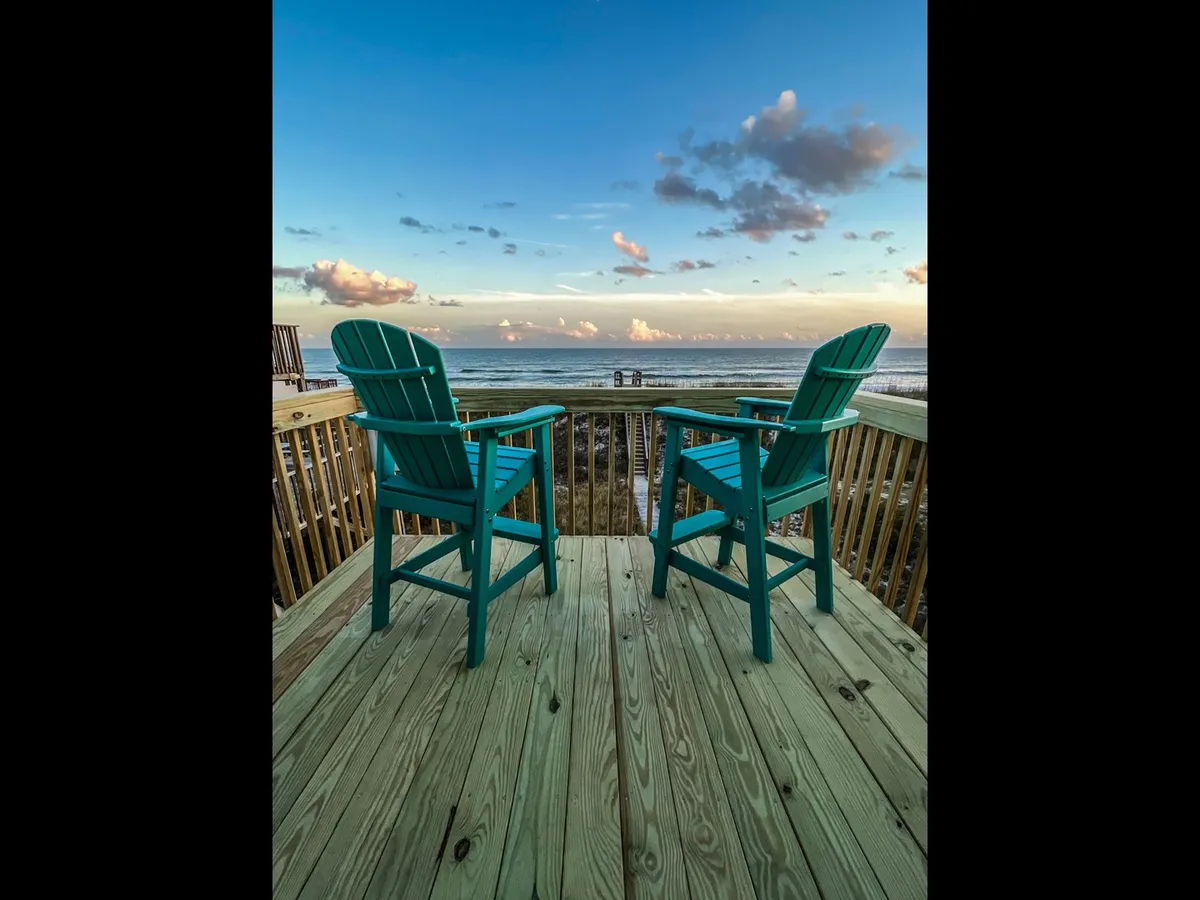 This exterior shot shows two teal Adirondack chairs placed on a deck overlooking the expansive ocean, with clouds dotting the sky. The scene suggests a tranquil outdoor space perfect for relaxing, socializing, or sharing a romantic moment while enjoying sweeping ocean views and gentle sea breezes.