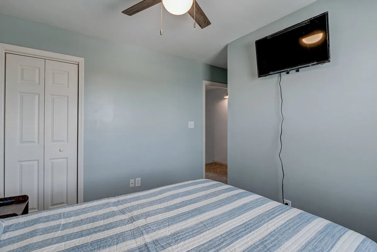 A well-lit bedroom with a striped bedding set, mounted TV, and ceiling fan. The neatly arranged bed and soft lighting evoke a sense of comfort and relaxation, while the TV and fan suggest modern amenities for guest convenience.