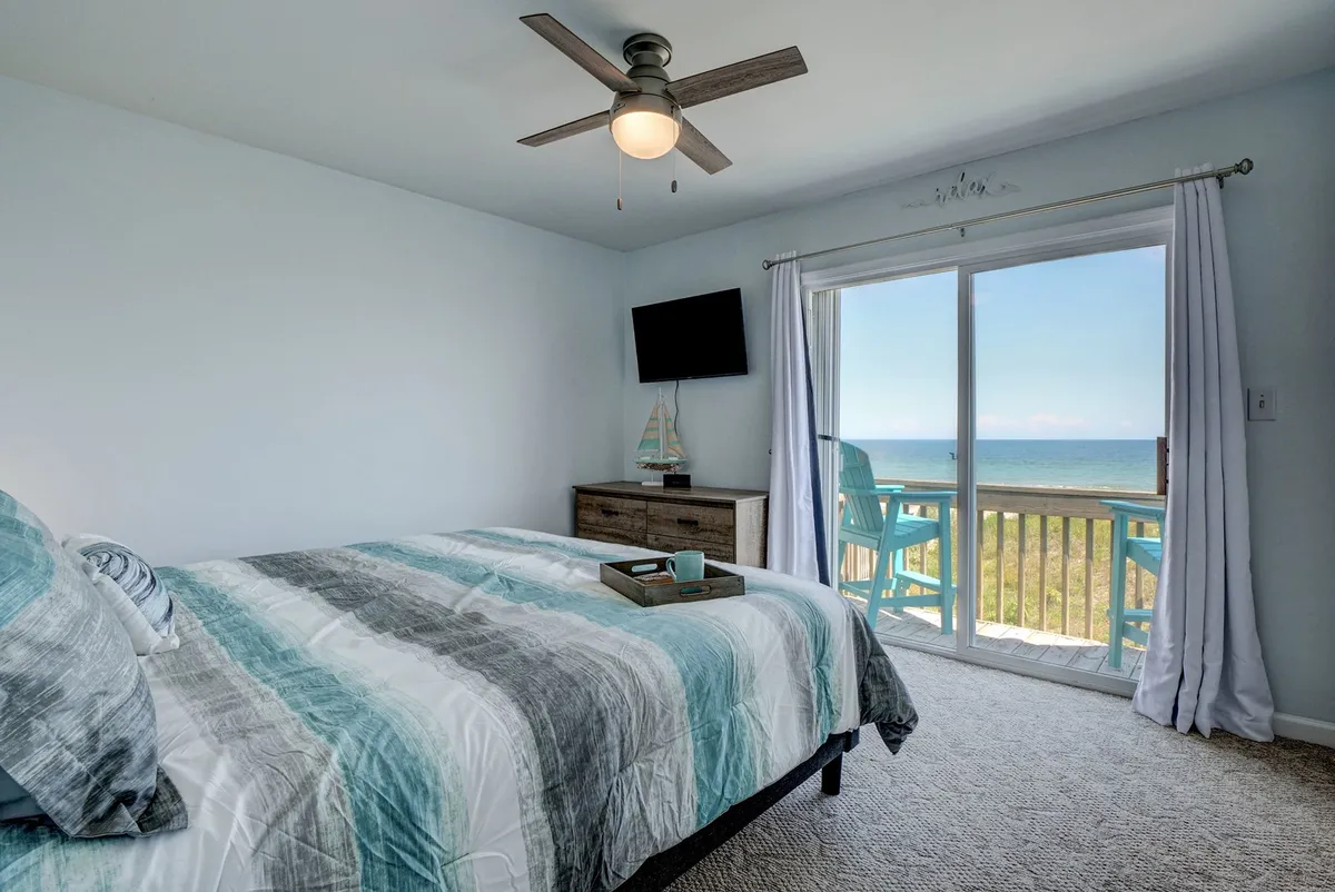 A tranquil bedroom featuring a bed with striped bedding positioned near a sliding glass door that opens to a private deck. The window reveals stunning ocean views, and a flat screen TV is mounted for convenience. Morning light streams in, creating an inviting atmosphere for relaxation or enjoying a sunrise coffee.