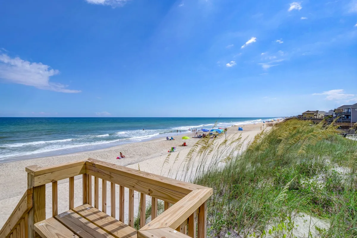 A panoramic view of the beach and ocean, with dune grass, wooden decking, and people enjoying various activities like picnicking, walking, and playing games along the shore.