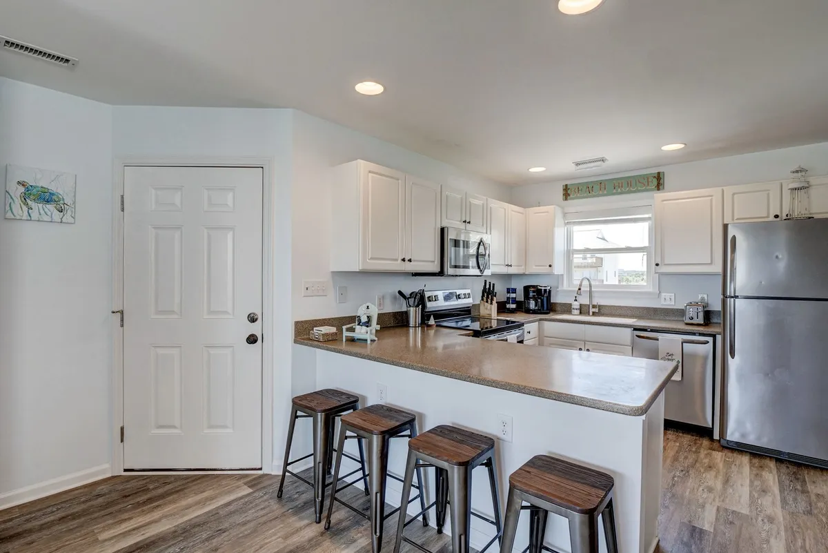 The image highlights a modern kitchen with a spacious island, stainless steel appliances, bar stools, and abundant natural light streaming in from a nearby window. The setup encourages shared cooking, casual breakfasts, or socializing around the island, perfectly illustrating the 'fully equipped kitchen' and the breakfast bar for sunset cocktails mentioned in the narrative.